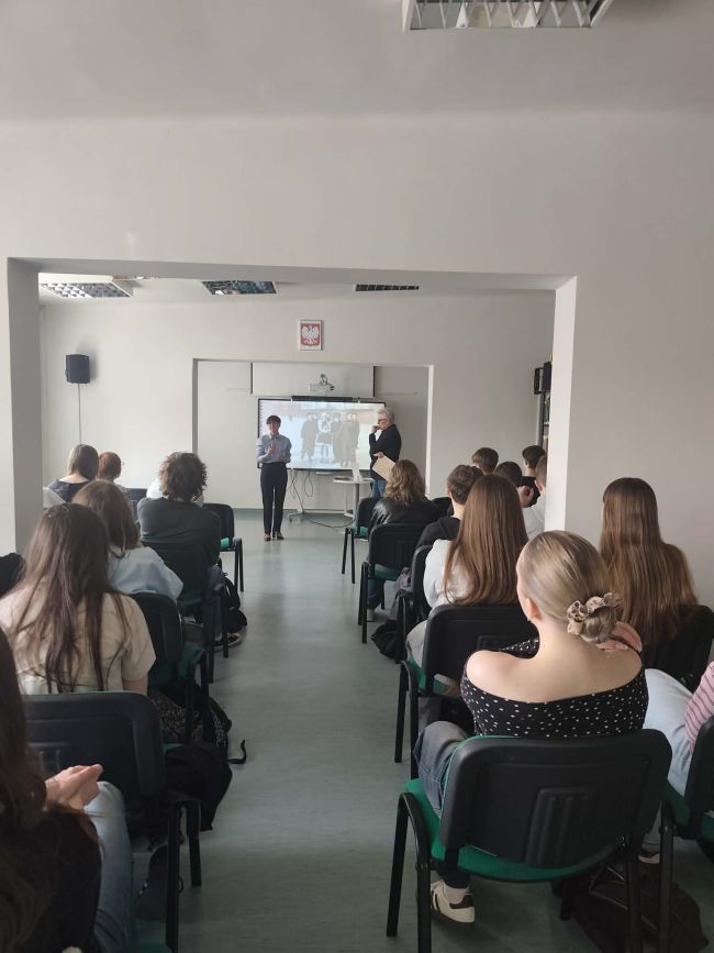 uczniowie na krzesłach słuchają wypowiedzi mężczyzny