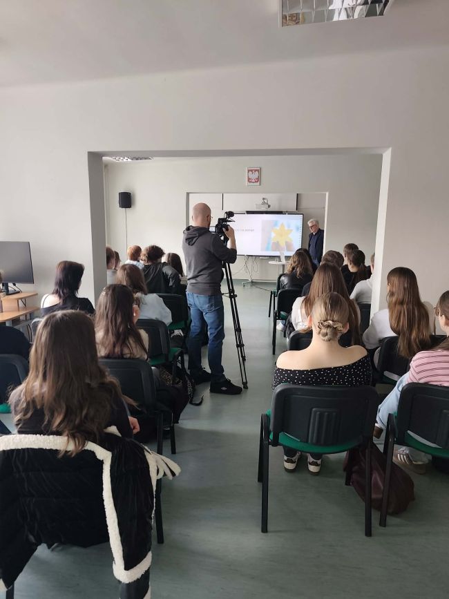 na pierwszym planie głowy, na drugim mężczyzna pokazujący coś na tablicy