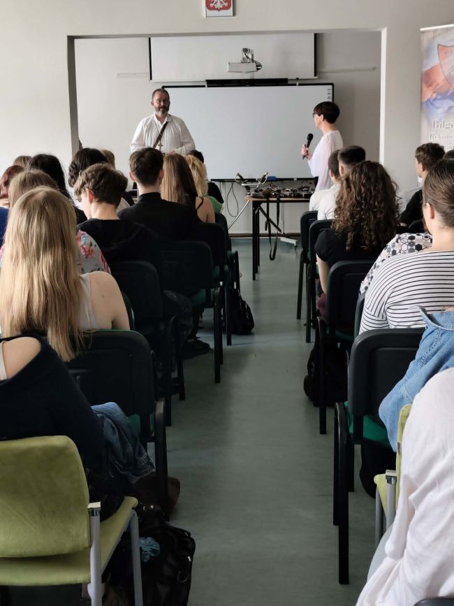 sala pełna ludzi, kobieta mówi do mikrofonu obok stoi mężczyzna w stroju powstańca