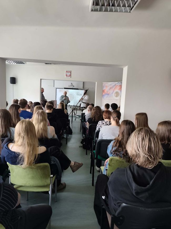 napis na tablicy multimedialnej, a przed nią leżą na stoliku militaria
