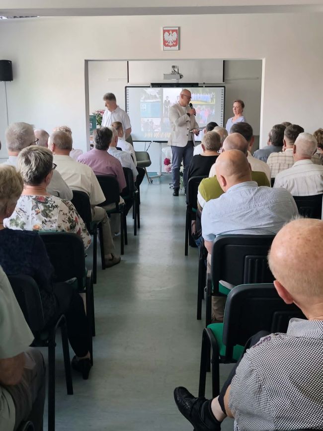 na pierwszym planie osoby siedzące na krzesłach, w tle osoby stojące pod tablicą multimedialną