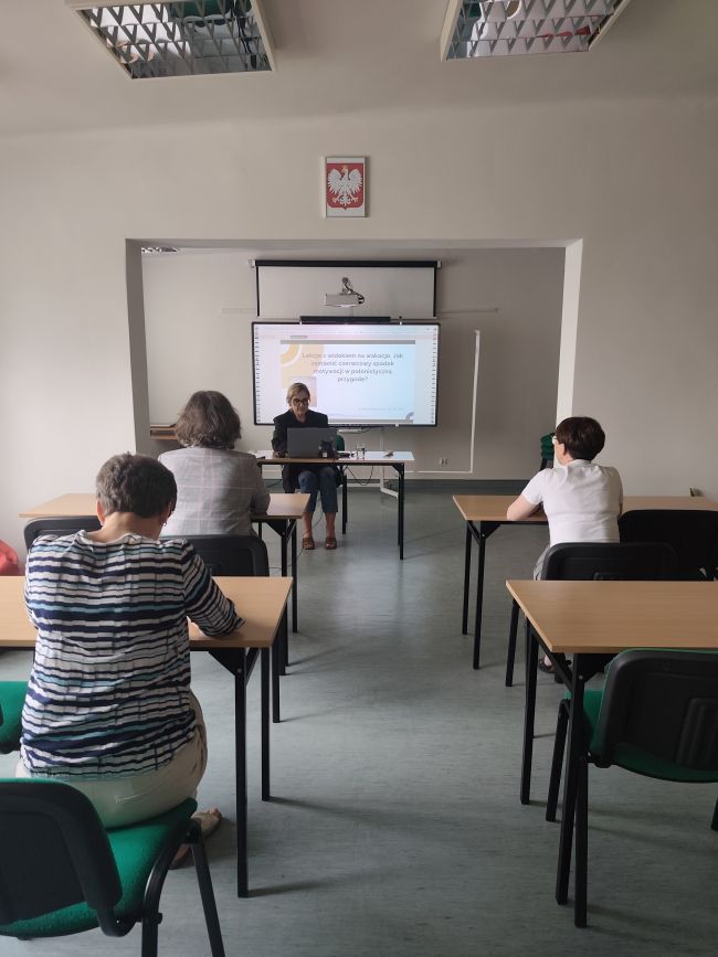 Osoby siedzące w ławkach, patrzące na tablicę multimedialną przed która siedzi kobieta prowadząca szkolenie