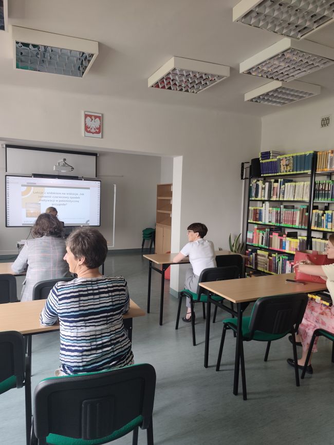 Osoby siedzące w ławkach, patrzące na tablicę multimedialną przed która siedzi kobieta prowadząca szkolenie