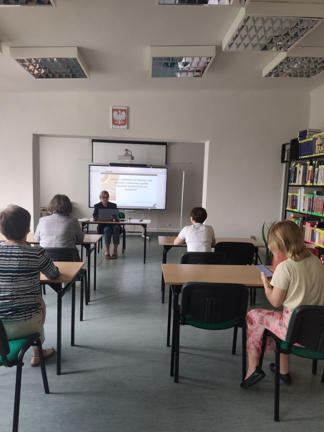 Osoby siedzące w ławkach, patrzące na tablicę multimedialną przed która siedzi kobieta prowadząca szkolenie