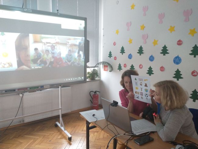 Biblioteka. Przy stoliku prowadzący zajęcia on-line. W tle tablica interaktywna z z widokiem prowadzących oraz grupy przedszkolaków