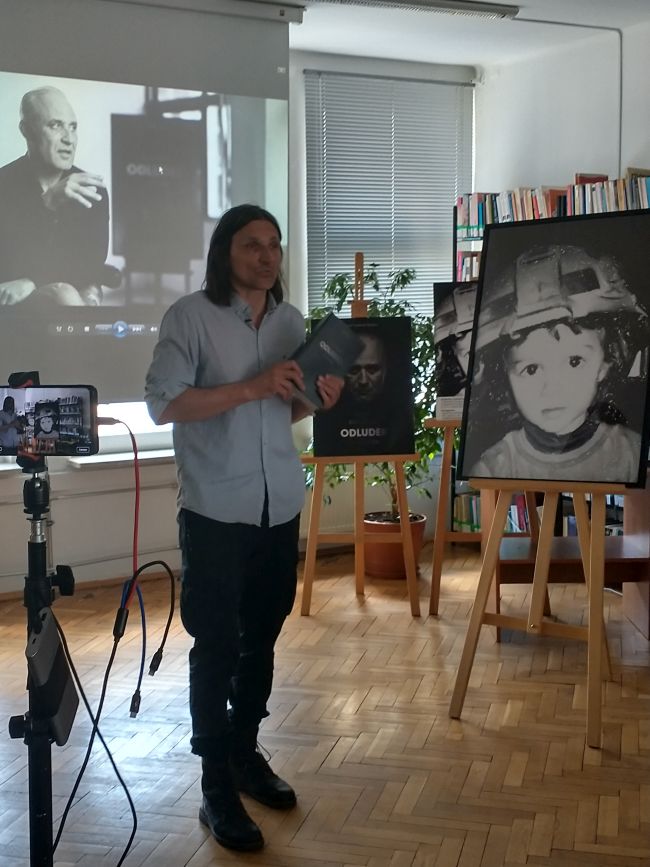 Na pierwszym planie Robert Kulik podczas spotkania autorskiego w bibliotece, prezentujący swoją debiutancką książkę. Obok na sztaludze jedna z rycin pochodząca z książki. W tle projekcja wywiadu z Przemysławem Bluszczem.