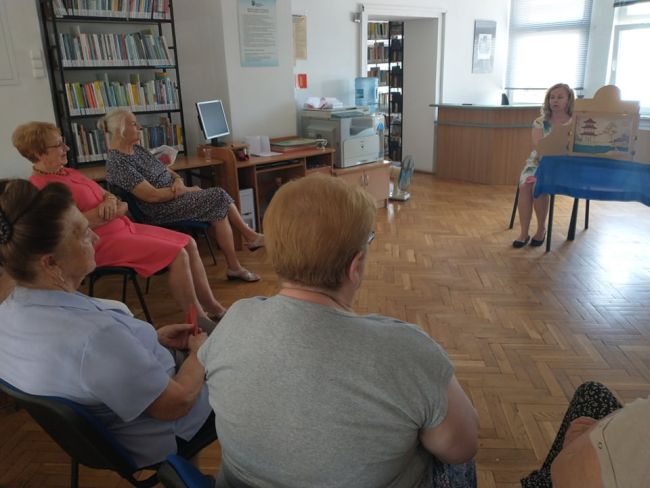 Biblioteka. Uczestnicy zajęć siedząc w półkolu słuchają historii czytanej przez nauczyciela bibliotekarza