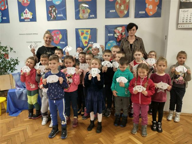 Biblioteka. Dzieci wraz z opiekunem i prowadzącą zajęcia pozują do wspólnej, pamiątkowej fotografii. W rękach trzymają wykonane przez siebie prace metodą origami.