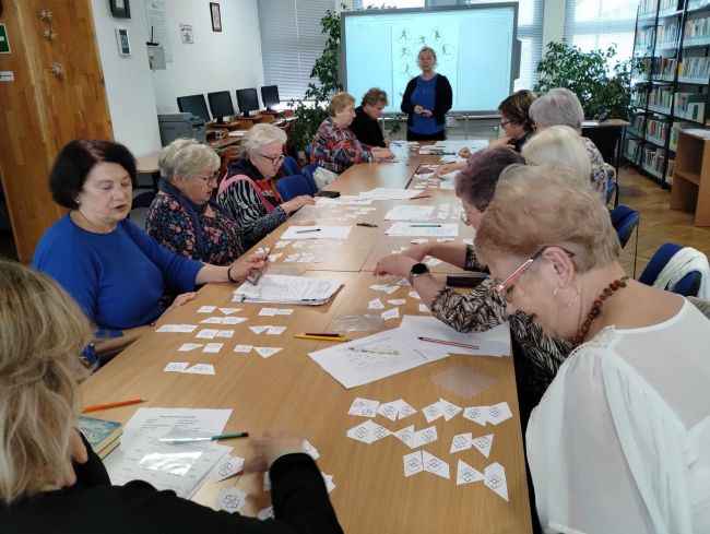 Seniorzy siedzą przy stołach, rozwiązują zadania. Prowadzące stoją przy tablicy interaktywnej.