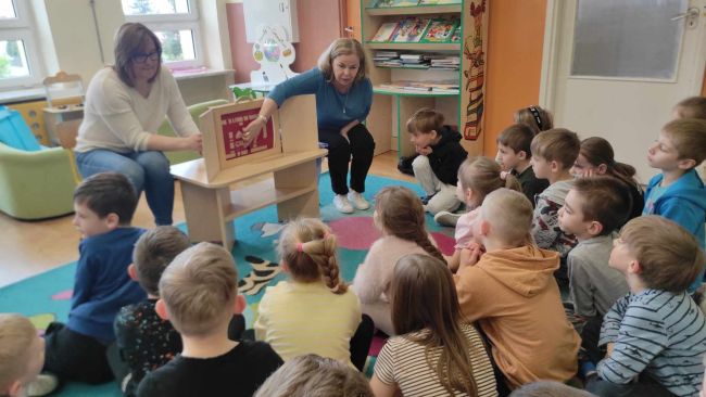 Dzieci siedzą na podłodze. Oglądają teatr Kamishibai. Prowadzące czytają historię