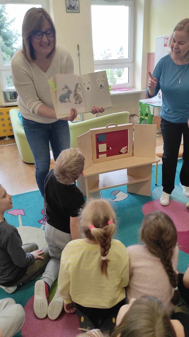 Dzieci siedzą na podłodze. Nauczycielki bibliotekarki prowadza zabawę pt. „oczami wyobraźni. Dzieci odgadują ukryte w książce obrazki.