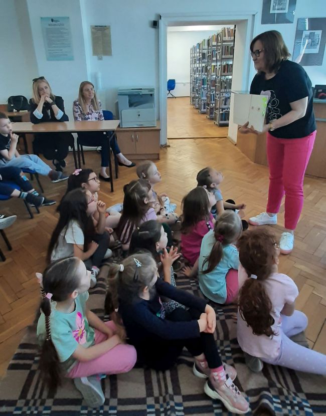 Prowadzące czytają dzieciom historię z wykorzystaniem teatrzyku Kamishibai.
Przedszkolaki siedzą na podłodze i słuchają.