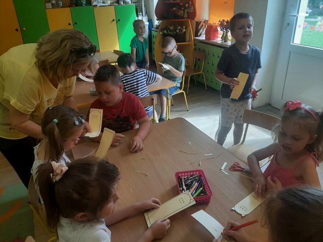 Nauczyciel pomaga dzieciom siedzącym przy stolikach wycinać zakładki
