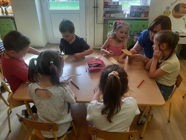 Przedszkolaki siedzą przy stolikach i malują, nauczyciel pomaga wycinać prace