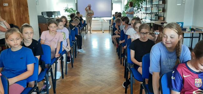 Uczniowie siedzą w rzędach na krzesłach, nauczyciele prowadzą zabawę.