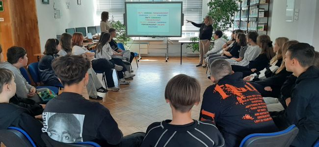 Prowadzące stoją przy tablicy interaktywnej. Uczniowie siedzą na krzesłach w kręgu. Wykonują zadanie