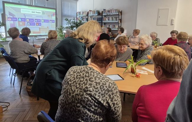 Uczestniczka rozwiązują Quiz w aplikacji Kahoot, wyświetlany na tablicy interaktywnej. Wykorzystują tablety. Prowadząca pomaga uczestniczkom.