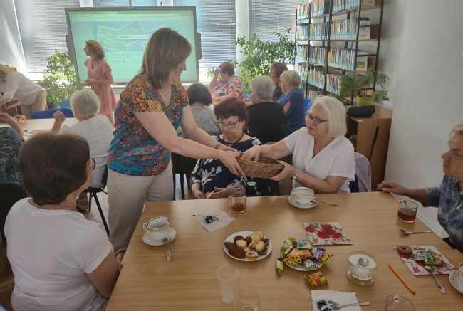 Słuchacze siedzą przy stolikach. Prowadząca rozdaje seniorkom koperty.