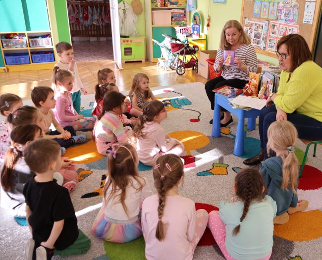 Nauczyciele siedzą na krzesłach i prowadzą zajęcia, dzieci  siedzą na dywanie i słuchają.