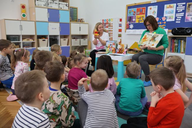 Nauczyciele czytają książkę dzieciom, które siedzą na dywanie i słuchają.