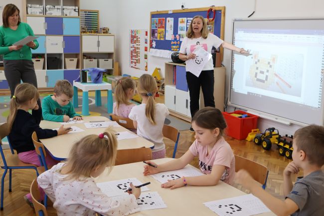 Dzieci siedzą przy stolikach i wykonują pracę z kodowania buzi Kubusia Puchatka, nauczyciel tłumaczy zadanie przy tablicy interaktywnej.