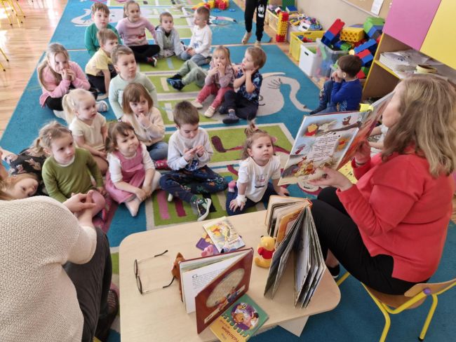 Nauczyciele czytają książkę, dzieci siedzą na dywanie i słuchają.