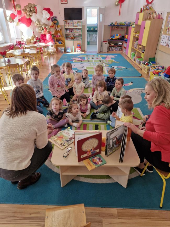 Nauczyciele prowadzą zajęcia, przedszkolaki siedzą na dywanie i słuchają.