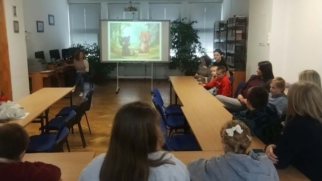 Prowadzące zajęcia siedzą i czytają bajkę, która wyświetlana jest na tablicy, dzieci wraz z opiekunami siedzą przy stolikach i słuchają opowieści.