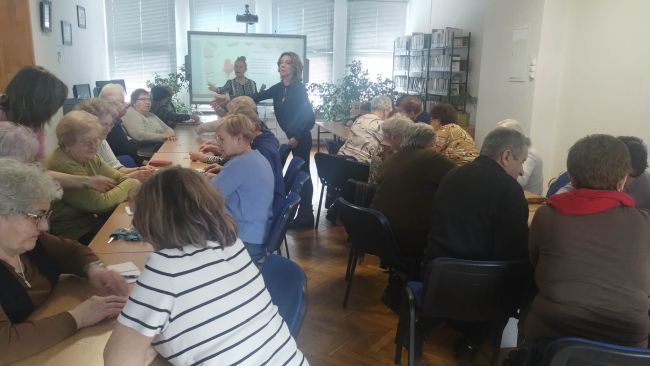 Słuchacze Uniwersytetu Trzeciego Wieku z Tomaszowa Lubelskiego siedzą przy stolikach, uczestniczą w zajęciach. Prowadzące tłumaczą wykonanie zadania.