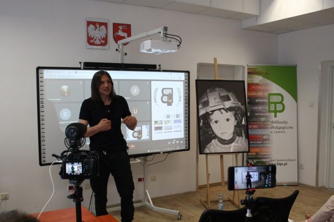 Na zdjęciu Robert Kulik opowiadający o swojej debiutanckiej książce podczas spotkania w Bibliotece Pedagogicznej w Zamościu. Autor jest ubrany na czarno (t-shirt i spodnie), ma czarne włosy do ramion.