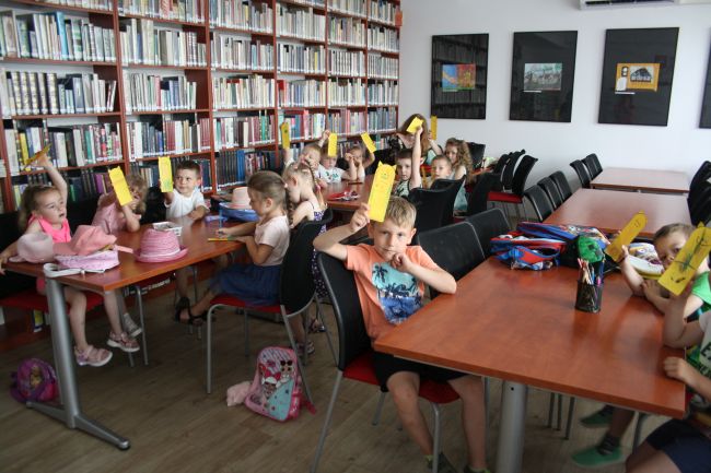 Uczestnicy zajęć siedzący przy stolikach i trzymający podniesione do góry wykonane przez siebie zakładkami do książek.