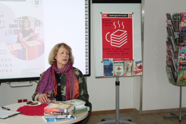 Na zdjęciu widać pisarkę Teresę Monikę Rudzką siedzącą przy stoliku, na którym leżą książki jej autorstwa. W rogu po prawej stronie na tablicy magnetycznej jest przyczepiony plakat Dyskusyjnego Klub Książki.
