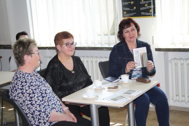 Na zdjęciu widać osoby uczestniczące w spotkaniu autorskim. Jedna z nich trzyma w ręku książkę 