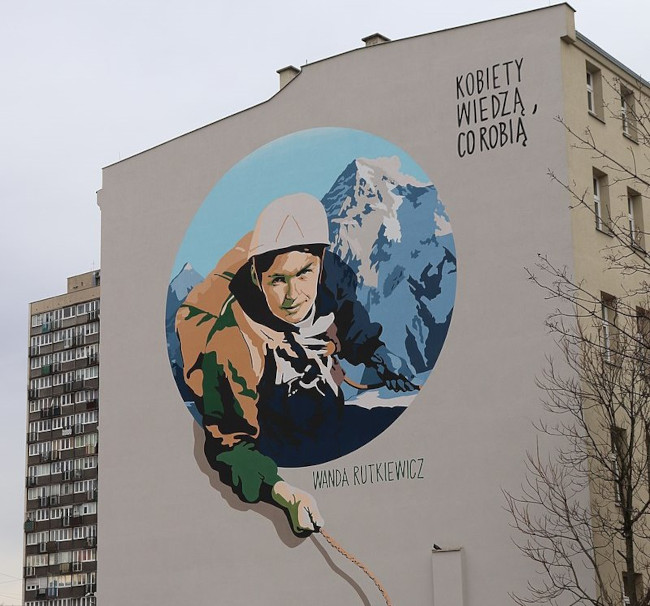 Zdjęcie prezentuje mural Wandy Rutkiewicz znajdujący się przy Placu Legionów we Wrocławiu. Mural jest wykonany na bloku mieszkalnym. Widać na nim Wandę Rutkiewicz w ubraniu górskim, trzymająca linę w ręku, a w tle widać ośnieżony szczyt. Nad wizerunkiem Wandy napis: 