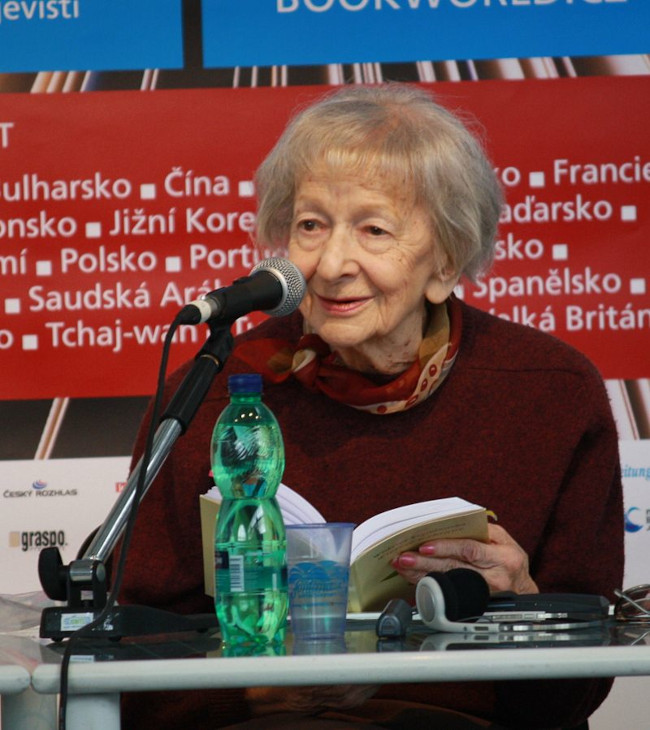 Zdjęcie przedstawia Wisławę Szymborską w 2010 roku, siedzącą przed mikrofonem.