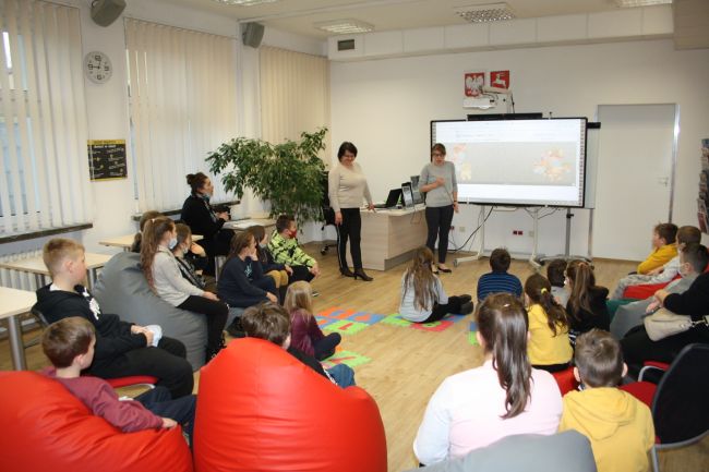 Początek zajęć o Kubusiu Puchatku w czytelni Biblioteki Pedagogicznej. W centralnej części stoją osoby prowadzące Barbara Mazurek i Agnieszka Kowalczuk, po prawej stronie jest tablica interaktywna, a po lewej duża roślina fikus. Przysłuchują im się uczestnicy, którzy siedzą na dużych kolorowych pufach oraz na piankowych puzzlach.