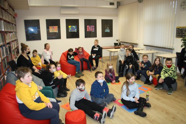 Na zdjęciu widać uczestników zajęć, którzy siedzą na kolorowych pufach oraz na piankowych puzzlach na podłodze i z zaciekawieniem słuchają. Na zdjęciu jest około 20 osób.
