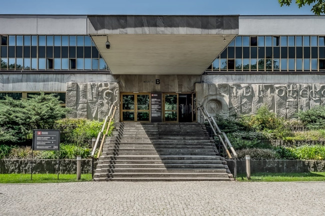 Na zdjęciu ukazane jest wejście do budynku Biblioteki Narodowej. Widać schody, otwarte drzwi, u góry rząd okiem. Po jednej i drugiej stronie schodów znajdują się różne rośliny.