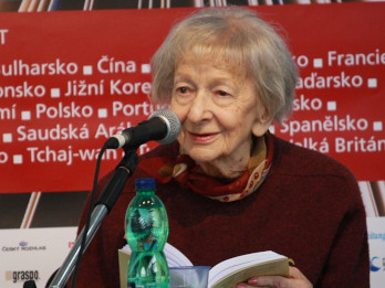 Zdjęcie przedstawia Wisławę Szymborską podczas spotkania autorskiego. Siedzi za stołem, w ręku trzyma książkę, mówi do mikrofonu. Na stole ustawiona jest butelka wody.