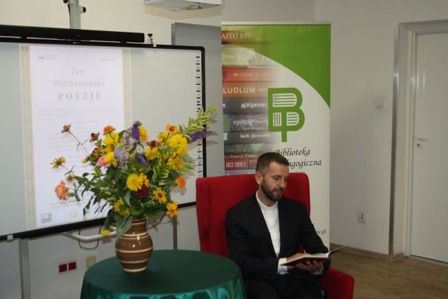 Dyrektor Katolickiej Szkoły Podstawowej ksiądz Mariusz Skakuj czyta poezję Jana Kochanowskiego. Siedzi przy stoliku, na którym w dużym wazonie jest bukiet złożony z wielu kwiatów.