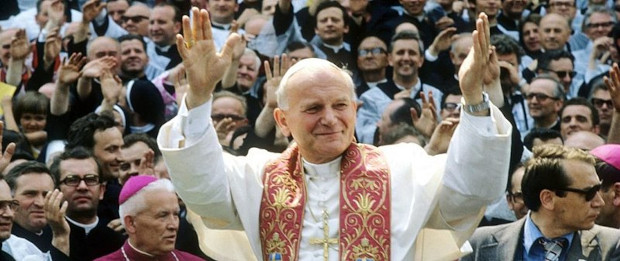 Zdjęcie przedstawia Jana Pawła II podczas pierwszej pielgrzymki do Polski w 1979 roku. Papież ma rozłożone do góry ręce, w tle za nim widać tłum ludzi.