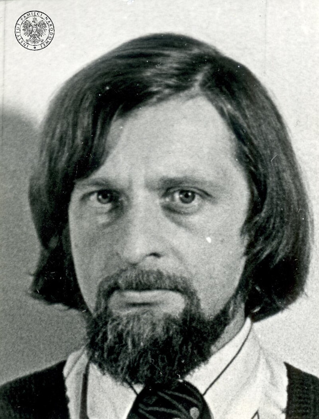 Fotografia przedstawia Janusza Andrzeja Zajdla. Uchwycone jest popiersie pisarza. Ma nieco dłuższe włosy zakrywające uszy. Ma krótką brodę i wąsy. Widać kawałek krawatu, białą koszulę oraz sweter. Zdjęcie pochodzi z akt paszportowych z ok. 1977 roku.