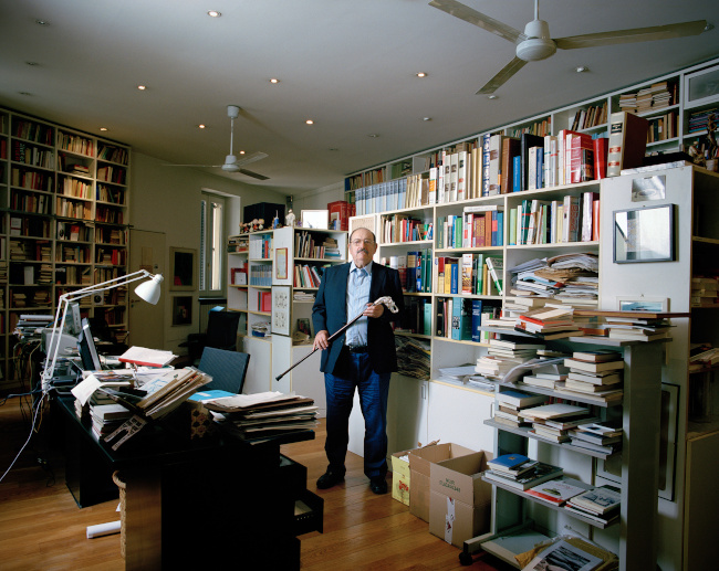 Zdjęcie przedstawia Umberto Eco w swoim domu na tle półek z książkami. Eco jest ubrany w marynarkę, koszulę oraz spodnie dżinsowe. Na nosie ma okulary. Po lewej stronie widać biurko z dużą lampką zaczepioną do biurka.