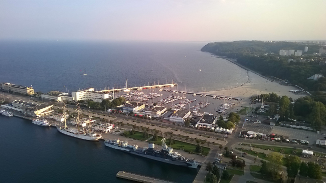 Zdjęcie Gdyni Śródmieście ukazanej od mola południowego. Na zdjęciu widać troszkę zabudowań miasta, a także morze, przycumowane statki oraz pływające łódki.