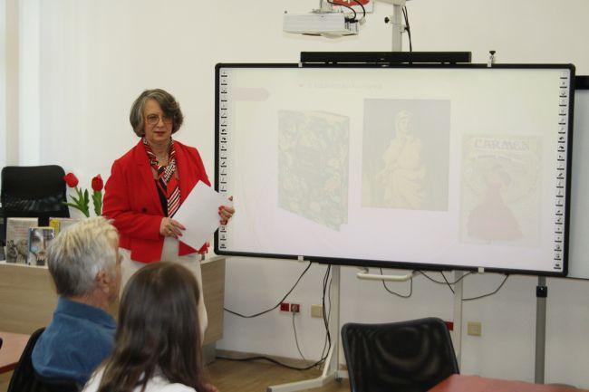 Dyrektor biblioteki dr Dorota Mach wygłasza prelekcję na temat kobiet w literaturze i malarstwie, której przysłuchują się seniorzy zebrani na kolejnym spotkaniu z cyklu