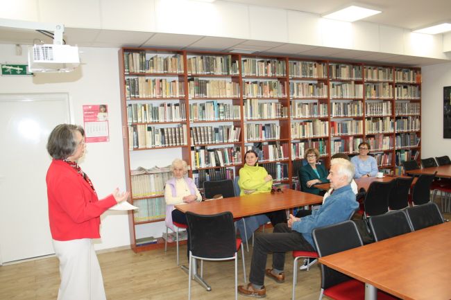 Dyrektor biblioteki dr Dorota Mach wygłasza prelekcję na temat kobiet w literaturze i malarstwie, której przysłuchują się seniorzy zebrani na kolejnym spotkaniu z cyklu