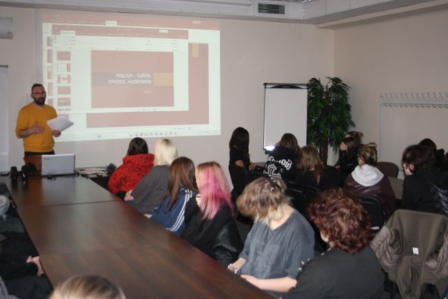 Łukasz Kot opowiada o pracy dziennikarza, młodzież siedząca przy stołach przysłuchuje się mu.